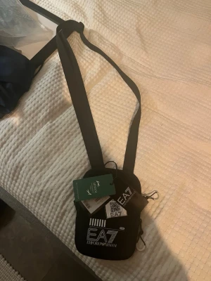 Svart axelväska EA7 Emporio Armani - Snygg svart axelväska från EA7 Emporio Armani med justerbar rem och vita logotexter både fram och bak. Väskan är kompakt och har dragkedja samt flera fack för småprylar. Tillverkad i slitstarkt syntetmaterial och perfekt för att bära det viktigaste.