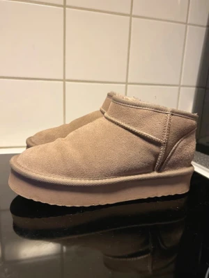 Beige fluffiga slip-in tofflor - Mysiga beige uggs med mjuk, fluffig insida och ovansida i mockaimitation. De har en tjock sula och en enkel, stilren. Uggsen  är ankelhöga och ger en varm och skön känsla.