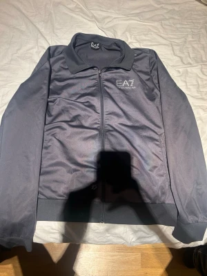 Svart jacka från EA7 Emporio Armani - Snygg grå hoodie från Emporio Armani EA7 med dragkedja framtill och diskret logga på bröstet. Klassisk design med ribbade muddar och mjukt material som känns skönt mot huden. Perfekt för dig som gillar stilrena och exklusiva plagg.