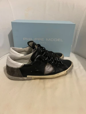 Philippe Model svarta sneakers - Svarta Philippe Model sneakers i läder med vita och grå detaljer. Klassisk låg modell med vita sulor och logotyp på sidan. Skorna har svarta snören och en unik textdetalj på plösen. Perfekta för en trendig och avslappnad stil.