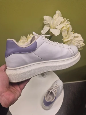Nelly shoes vita chunky sneakers med lila detalj 38 - Stilrena vita sneakers med lila detalj vid hälen och tjock sula. Klassisk design med snörning och diskreta hål på sidan för ventilation. Tillverkade i slätt syntetmaterial som ger en clean look. Perfekta för dig som gillar minimalistisk stil.