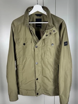 Vindjacka från J.Lindeberg - Stilren olivgrön vindjacka från J.Lindeberg med tryckknappar framtill, två bröstfickor och hög krage. Jackan har lång ärm och diskret logotyp på ärmen. Perfekt för dig som gillar minimalistisk design och funktion.