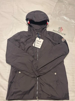 Moncler Vindjacka - Perfekt skick! - Snygg vindjacka från Moncler med huva och dragkedja framtill. Jackan har Moncler-logga på ärmen, två fickor med tryckknappar och detaljer i rött, vitt och blått på huvan. Tillverkad i lätt och slitstarkt material, perfekt för blåsiga dagar. Fick denna som gåva men tyvärr så vart den liten på mig så den måste tyvärr säljas.