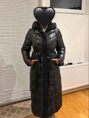 Lång svart dunjacka med huva - Snygg lång svart dunjacka med bälte i midjan och stor huva. Jackan har quiltad design, dragkedja framtill och två fickor på sidorna. Perfekt för kalla vinterdagar och ger en stilren siluett. Använd två gånger 🖤
