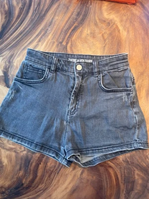 Gråa high waist shorts Zadig & Voltaire - Snygga gråa denimshorts från Zadig & Voltaire med hög midja och klassisk femficksdesign. Shortsen har en knapp och dragkedja framtill samt broderad ZV-logga på ena bakfickan. Perfekta för sommardagar och enkel att matcha med olika toppar.