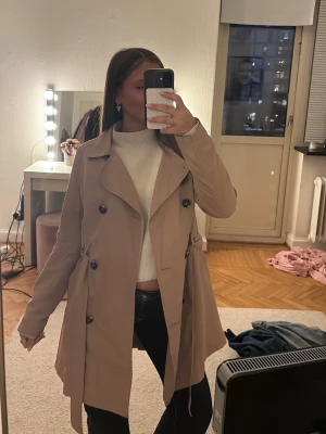 Beige trenchcoat med knappar - Stilren beige trenchcoat med dubbelknäppning och klassisk krage. Jackan har ett bälte i midjan och svarta knappar framtill. Perfekt för lager-på-lager och ger en snygg siluett. Passar till många olika outfits.