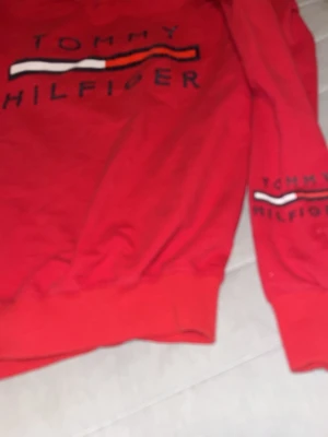 Röd Tommy Hilfiger hoodie - Snygg röd hoodie från Tommy Hilfiger med stort tryck på bröstet och ärmen. Klassisk logga i vitt, rött och marinblått. Hoodien har långa ärmar och ribbade muddar. Perfekt för dig som vill ha en stilren och bekväm look.