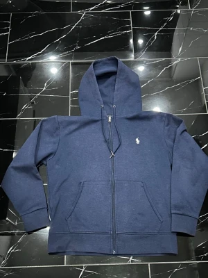 Marinblå zip hoodie från Ralph Lauren - Klassisk marinblå hoodie med dragkedja från Polo Ralph Lauren. Hoodien har huva med dragsko, två fickor framtill och den ikoniska vita polospelaren broderad på bröstet. Tillverkad i mjuk bomullsblandning som är perfekt för chill dagar.