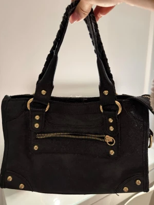 Svart handväska med gulddetaljer - Stilren svart handväska med dubbla handtag och snygga guldfärgade nitar och dragkedja framtill. Väskan har en rektangulär form och är tillverkad i ett tygmaterial. Perfekt för dig som gillar en edgy look och vill ha plats för det viktigaste.