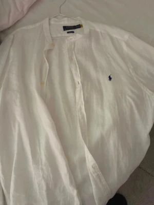 Vit linneskjorta från Polo Ralph Lauren - Klassisk vit skjorta i linne från Polo Ralph Lauren med den ikoniska broderade loggan på bröstet. Skjortan har knappar framtill och en avslappnad passform, perfekt för varma dagar. Enkel och stilren design som funkar till mycket.