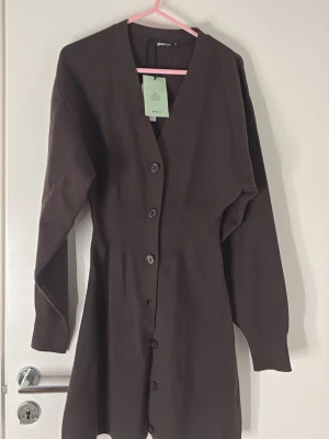 Brun lång kofta från Gina Tricot - Mörkbrun lång kofta med knappar hela vägen framtill från Gina Tricot. Koftan har v-ringning, långa ärmar och markerad midja för en snygg siluett. Perfekt att slänga över en outfit för extra stilpoäng.