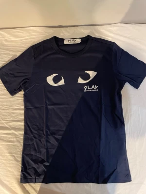 Mörkblå Comme des Garçons PLAY t-shirt - Snygg mörkblå t-shirt från Comme des Garçons PLAY med ikoniska ögon-trycket och logga på bröstet. Klassisk passform med rund hals och korta ärmar. Tillverkad i mjuk bomull som känns skön mot huden.