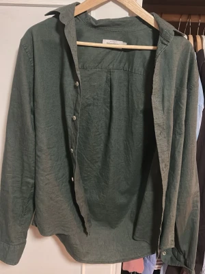 Grön linneskjorta från Dressman - Stilren grön skjorta från Dressman i mjukt linne. Klassisk krage, långa ärmar med manschettknappar och knäppning framtill. Perfekt för en avslappnad men snygg look. Skjortan har en loose passform och enkel design utan mönster.