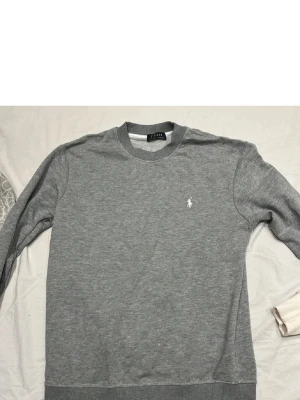 Grå sweatshirt från Polo Ralph Lauren - Klassisk grå sweatshirt från Polo Ralph Lauren med rund halsringning och diskret broderad logga i vitt på bröstet. Tröjan har ribbade muddar vid ärmslut och nederkant. Perfekt för en avslappnad och stilren look.