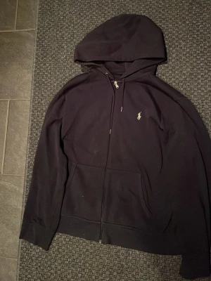 Svart hoodie från Ralph Lauren - Säljer en svart hoodie från Ralph Lauren med dragkedja framtill och klassisk vit logga på bröstet. Hoodien har huva med dragsko och två stora fickor framtill. Tillverkad i mjukt bomullsmaterial, perfekt för en avslappnad och stilren look.