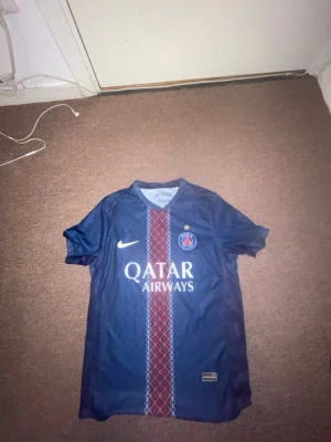 PSG matchtröja blå Nike - Snygg blå PSG matchtröja från Nike med klubbmärke och Qatar Airways-tryck på bröstet. Tröjan har ett rött och vitt mönster längs mitten och V-ringad hals. Perfekt för dig som älskar fotboll och vill representera Paris Saint-Germain.