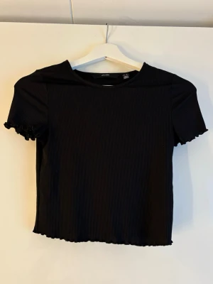 Svart ribbad croppad topp - En svart ribbad croppad topp med korta ärmar och vågig kant vid ärmslut och nederkant. Enkel och stilren design som passar perfekt till jeans eller kjol. Gjord i mjukt material för en bekväm känsla.