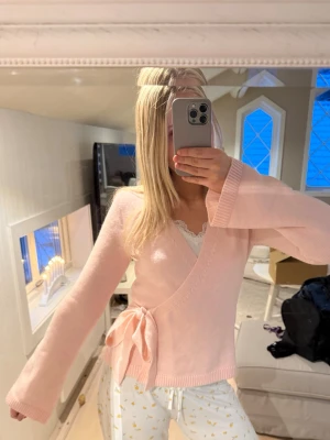 Ljusrosa omlottkofta med knytband - Supersöt ljusrosa omlottkofta med breda ärmar och knytband i midjan. Koftan har ribbade muddar och en mjuk, stickad känsla. Perfekt för dig som gillar en feminin och avslappnad stil.