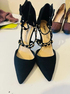 Svarta pumps med nitar från Jumex - Helt nya med prislapp Snygga svarta pumps från Jumex med spetsig tå och smala klackar. Skorna har flera remmar dekorerade med guldfärgade nitar och spänne vid ankeln. Materialet är mockaimitation som ger en lyxig känsla. Perfekta för dig som vill sticka ut med din stil.