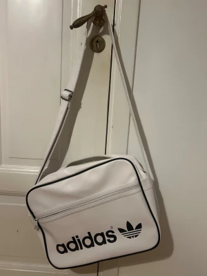 Vit axelväska från Adidas - Snygg vit axelväska från Adidas med svart logga och text på framsidan. Väskan har justerbar axelrem, svart piping längs kanterna och ett stort huvudfack samt en yttre ficka med dragkedja. Tillverkad i ett glansigt konstläder som ger en retro känsla.