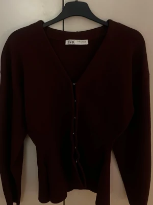 Vinröd kofta från Zara - Mörkröd kofta från Zara med v-ringning och knappar framtill. Långärmad modell i mjukt material som sitter snyggt på kroppen. Perfekt att slänga över en topp eller bära stängd för en clean look.