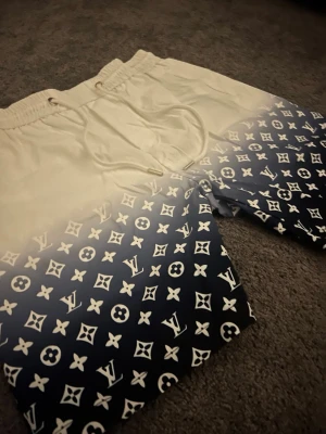 Vit-blå LV shorts med snörning - Säljer ett par snygga shorts från Louis Vuitton med beige ovandel och blå nederdel täckt av klassiskt LV-mönster. Shortsen har snörning i midjan och sidofickor. Perfekta för sommaren och ger en lyxig vibe till din outfit.