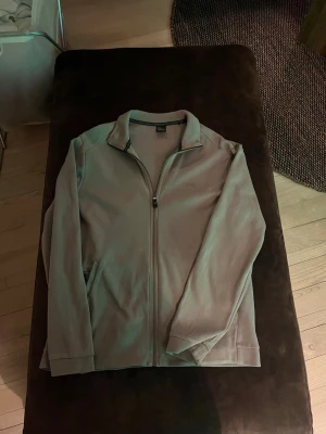 Grå ziptröja från BOSS - Säljer en stilren grå ziptröja från BOSS med hög krage och hel dragkedja framtill. Tröjan har diskret logga på bröstet och ribbade detaljer vid kragen. Perfekt för en avslappnad men snygg look.