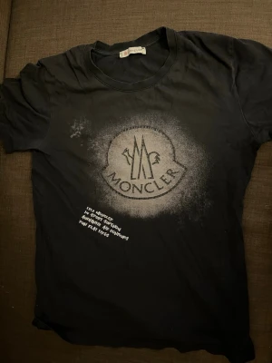 Svart Moncler t-shirt med tryck - Cool svart t-shirt från Moncler med stort logotryck framtill i vitt och spray-effekt runt loggan. Baksidan har ett stort Moncler-tryck i vitt. Klassisk rund hals och korta ärmar. Perfekt för dig som gillar streetwear och vill sticka ut.