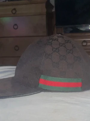 Gucci keps med GG-mönster - Säljer en svart keps från Gucci med klassiskt GG-monogram över hela kepsen och en grön-röd rand på sidan. Kepsen har böjd skärm och justerbart spänne bak i läder. Materialet är tyg och detaljerna ger en lyxig känsla. Perfekt för dig som gillar exklusiva accessoarer.