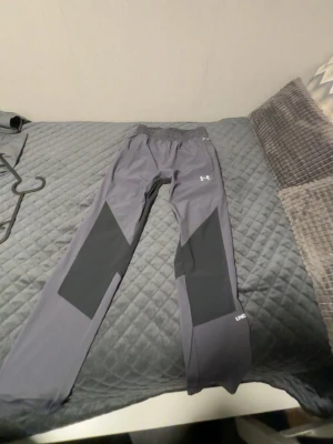 Grå träningsbyxor från Under Armour - Snygga grå träningsbyxor från Under Armour med svarta detaljer på benen. Byxorna har resår i midjan och sidofickor. Perfekta för gymmet eller löpning. Tillverkade i ett lätt och stretchigt funktionsmaterial.