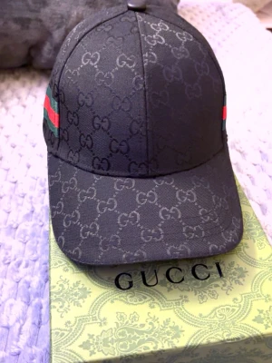 Svart keps från Gucci med GG-mönster - Säljer en svart keps från Gucci med det klassiska GG-monogrammet över hela kepsen. På sidorna finns Guccis ikoniska gröna och röda ränder. Justerbar rem baktill i svart läder med präglad logotyp. Perfekt för dig som gillar exklusiva accessoarer.