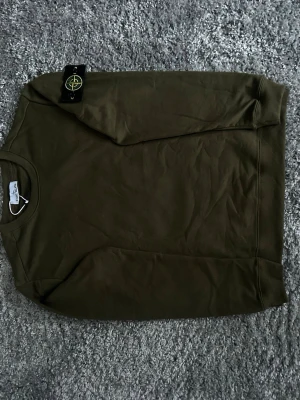 Olivgrön sweatshirt från Stone Island - Snygg olivgrön sweatshirt från Stone Island med klassisk rund hals och ikonisk logotyp-patch på ärmen. Tröjan är tillverkad i mjuk bomull och har ribbade muddar vid ärmslut och nederkant. Perfekt för en avslappnad streetwear-look.
