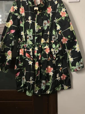 Jacka Blommig parkajacka från Baker - Snygg svart parkajacka från Baker med färgglatt blommönster och fåglar. Jackan har långa ärmar, dragkedja och knappar framtill samt justerbar midja. Perfekt för dig som vill sticka ut med en unik och färgstark look. I storlek 11 års ålder 