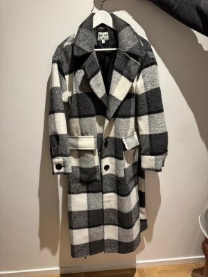 Rutig kappa från NA-KD - Snygg lång kappa från NA-KD i svartvitt rutigt mönster. Kappan har bred krage, knappar framtill, stora fickor och ett matchande knytskärp i midjan. Material som håller dig varm och trendig under kalla dagar.