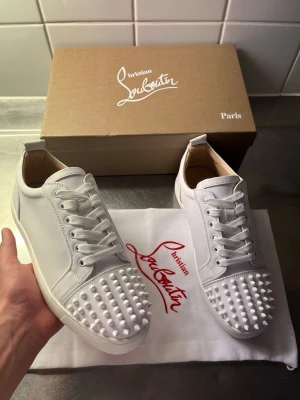 Vita Christian Louboutin sneakers med nitar - Säljer ett par vita sneakers från Christian Louboutin med ikoniska röda sulor och coola vita nitar på tån. Skorna är låga och har en clean design i läder med vita snören. Perfekta för dig som vill sticka ut med exklusiv stil.