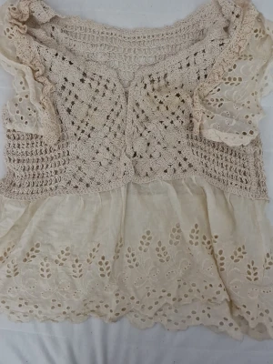 Beige virkad blus med spetsdetaljer - Supersöt beige blus med virkade partier upptill och broderade spetsdetaljer nertill. Blusen har korta ärmar med volang och ett romantiskt, bohemiskt uttryck. Perfekt till jeans eller kjol för en trendig look. PRIS KAN DISKUTERAS