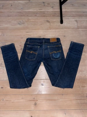 Mörkblå jeans från Nudie Jeans - Säljer ett par mörkblå jeans från Nudie Jeans med klassisk femficksmodell och raka ben. Jeansen har kontrastsömmar och Nudie-logga på bakfickan. Tillverkade i slitstark denim, perfekta för en avslappnad och stilren look. Sitter slim :)