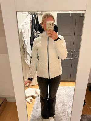 Rossignol skidjacka  - Vit vindjacka med svart foder i huvan och svarta detaljer vid dragkedja och ärmslut. Jackan har två fickor framtill, sportig passform och en liten logga på ärmen. Snygga svarta ränder på sidan ger en modern look. Perfekt för blåsiga dagar.