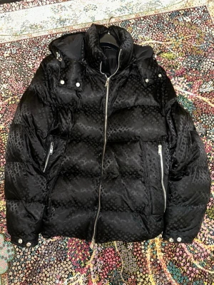 BOSS CORBINIAN1 - Vinterjacka - svart - Snygg svart pufferjacka från BOSS med diskret mönster i tyget. Jackan har hög krage, dragkedja framtill och två stora fickor med dragkedja. Stora silvriga knappar och detaljer vid axlarna ger en cool look. Perfekt för kalla vinterdagar.