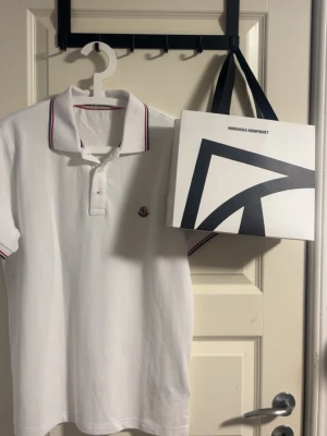 Moncler piketröja - Klassisk vit pikétröja från Moncler. Produkten är köpt från NK och har använts ett få antal gånger. NK påse ingår. Den är i väldigt bra skick utan några deffekter!
