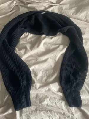 Svart ribbstickad bolero - Svart ribbstickad bolero med långa ärmar och bred mudd vid ärmsluten. Perfekt att slänga över en topp för extra värme och stil. Materialet känns mjukt och stretchigt, vilket gör den bekväm att bära.