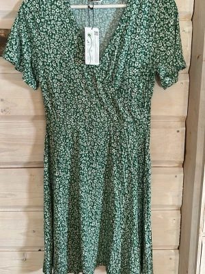 Grön blommig omlottklänning - Söt grön kortklänning med vitt blommönster och omlottdesign. Klänningen har v-ringning och korta ärmar, vilket ger en avslappnad och somrig känsla. Perfekt för dig som gillar en feminin och trendig stil.