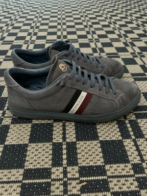 Grå Moncler sneakers i mocka - Snygga grå sneakers från Moncler i mjuk mocka med mörkblå snören och klassiska Moncler-ränder i svart, vitt och vinrött på sidan. Låg modell med rund tå och diskret logga på plösen. Perfekta för en clean och stilren look.