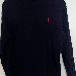 Mörkblå kabelstickad tröja Polo Ralph Lauren - Kabelstickad mörkblå tröja från Polo Ralph Lauren med klassisk röd logga broderad på bröstet. Tröjan har rund halsringning. Mycket bra skick!