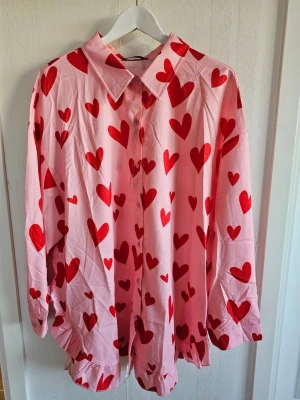 Rosa skjorta med röda hjärtan - Säljer en oversized rosa skjorta med röda hjärtan över hela plagget. Skjortan har klassisk krage, knäppning framtill och långa ärmar. Perfekt statement-piece för dig som vill sticka ut med ett lekfullt mönster. Materialet känns silkeslent. Storlek 4XL/EU54