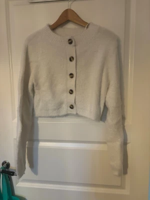 Vit fluffig cropped kofta - Mysig vit cropped kofta med stora bruna knappar framtill. Koftan har lång ärm, ribbstickad kant och är gjord i ett fluffigt material som känns riktigt mjukt. Perfekt för dig som gillar en enkel men trendig look.