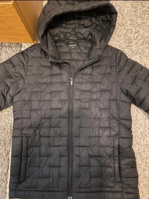 Svart pufferjacka med huva - Säljer en svart pufferjacka från Everest med huva och dragkedja framtill. Jackan har quiltat mönster och två sidofickor. Perfekt för kalla dagar och riktigt skön att bära. Materialet är lätt och isolerande.