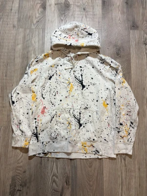 Abercrombie & Fitch - Säljer en vit hoodie med huva och känguruficka. Hoodien har ett unikt färgstänksmönster i svart, gult och rosa som ger en kreativ och trendig vibe. Tillverkad i mjukt bomullstyg och har ribbade muddar vid ärmar och nederkant. Perfekt för dig som gillar streetwear och vill sticka ut. Storlek XL Passar M/L