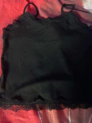 Svart spetslinnetopp med tunna band - Snygg svart linnetopp med tunna axelband och spetsdetaljer längs urringning och nederkant. Perfekt att styla lager på lager eller bära som den är. Materialet känns mjukt och följsamt.