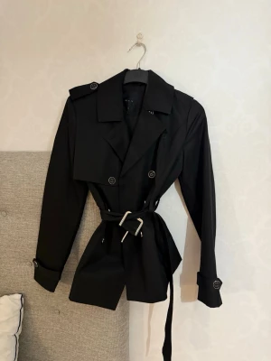 Svart trenchcoat med bälte från Vero Moda - Stilren svart trenchcoat från Vero Moda med klassisk dubbelknäppning, axelklaffar och avtagbart midjebälte. Jackan har långa ärmar med dekorativa knappar vid ärmsluten och en tidlös krage. Perfekt för dig som gillar minimalistisk och trendig stil.